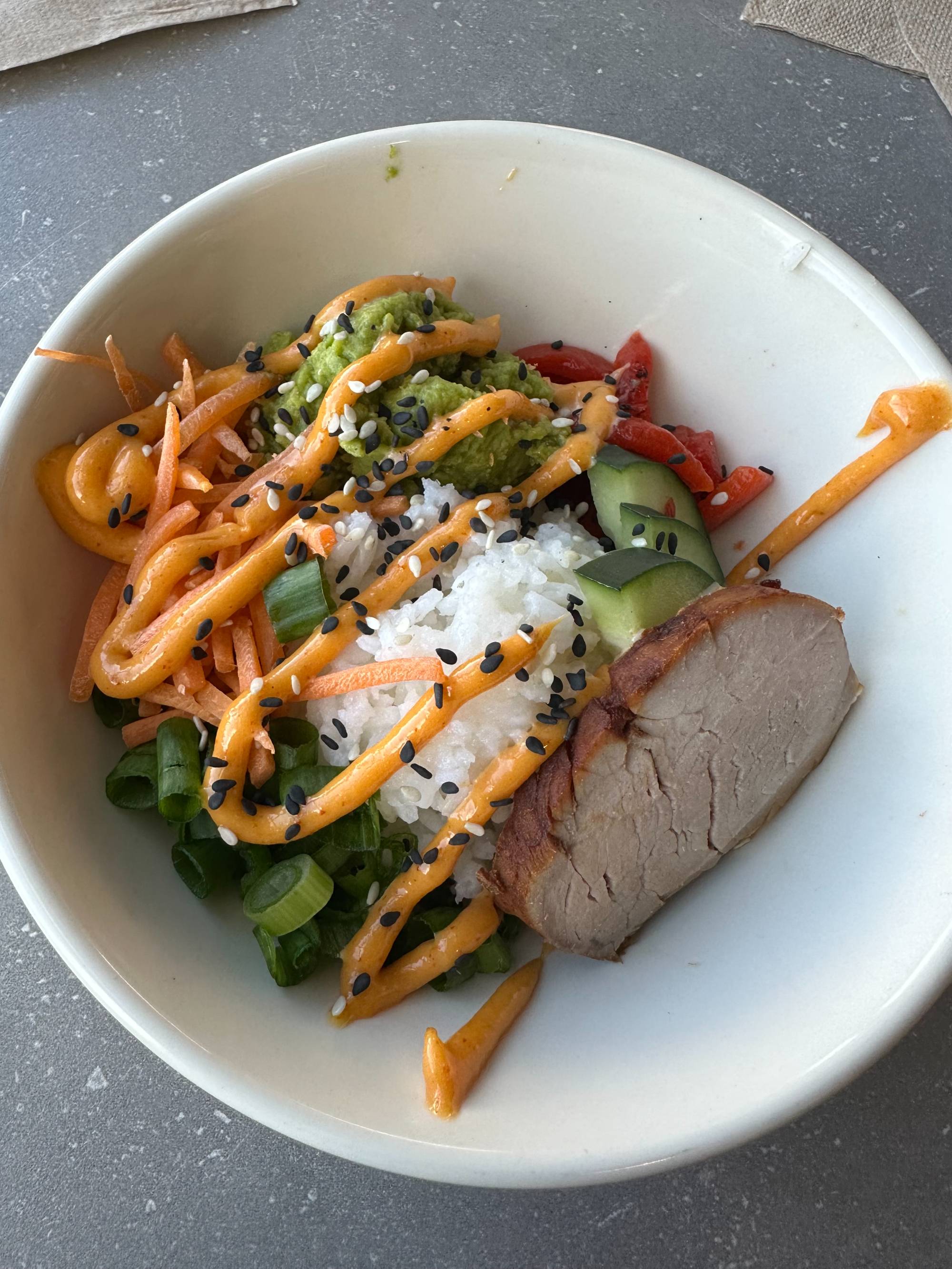 Colorful bowl with white rice, green onion, cucumber, avocado, pork, sesame seeds, and an orange sauce.